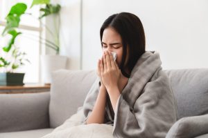 A sick Asian young woman sneezing into tissue paper while covered with a blanket and sitting on sofa in the living room - Sugar Land Mold Remediation.