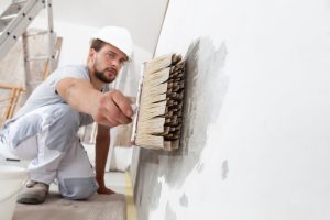construction worker painter man with protective helmet, brush in hand restores and paint the wall, inside the building site of a house, closeup - Painting over mold